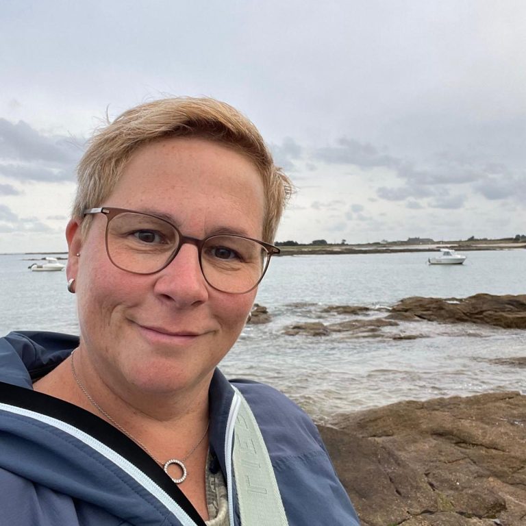 Person mit Brille lächelt am Strand bei bewölktem Wetter, Boote im Hintergrund.