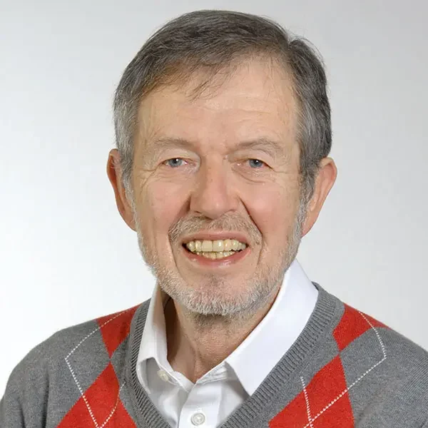 Älterer Mann mit grauem Haar, weißem Hemd und grauem Pullover mit roten Rauten.