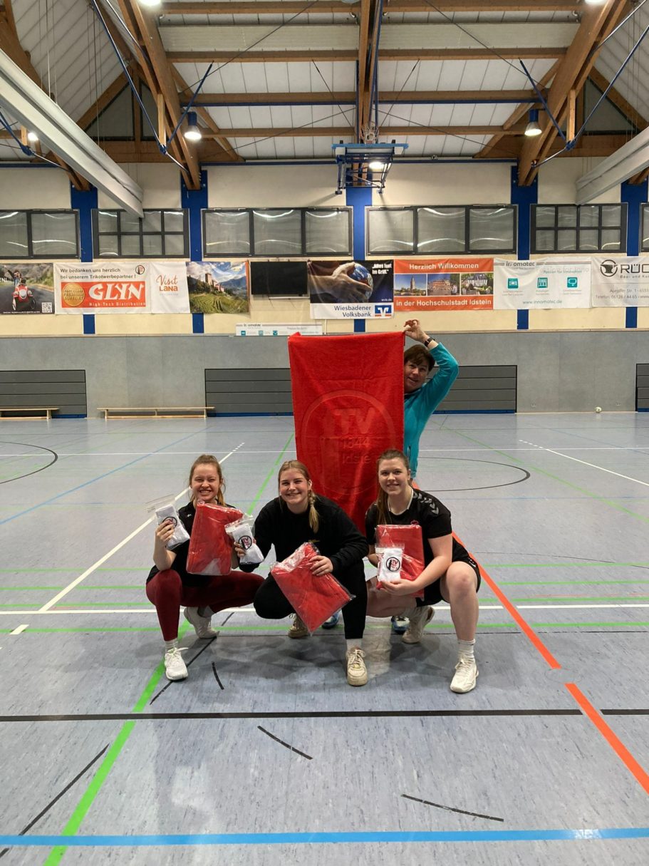 Drei Sportlerinnen sitzen auf dem Boden und halten rote Kissen und ein großes rotes Banner.